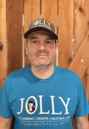 Man wearing a blue JOLLY Plumbing t-shirt and a dark cap, standing in front of a wooden fence.