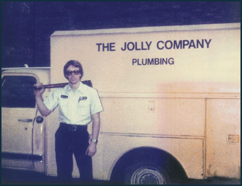 Plumber in uniform stands beside a service van labeled The Jolly Company Plumbing, with a wrench over his shoulder.