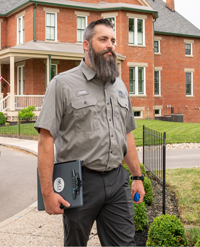 Bearded security guard in a gray uniform walks along a sidewalk outside a brick building, carrying a clipboard and pen.