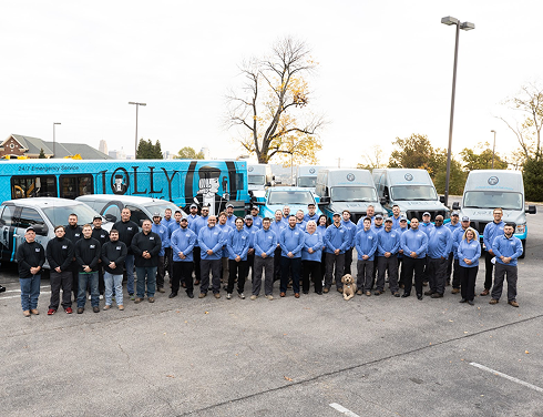 Group of employees in blue shirts and dark pants posing for a team photo in a parking lot, with a row of blue service vans behind and a dog at the front.