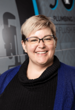 Smiling woman with short blonde hair and glasses, wearing a black turtleneck and blue cardigan, standing in front of a blue-gray wall.