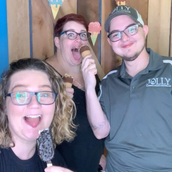 Three adults smiling and posing with ice cream treats in front of a wood-paneled wall.