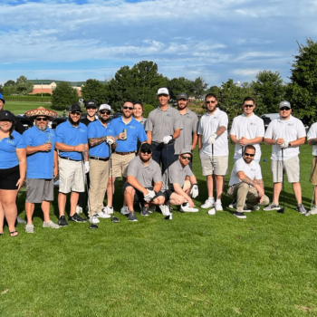 Group of golfers posing for a photo on a sunny golf course, many in blue shirts, with clubs in hand and a clubhouse visible in the background.