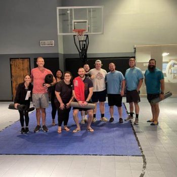 Group of adults standing on gym mats in a gym, posing with a basketball hoop behind them.