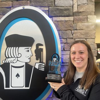 Smiling woman in a dark jacket holds a trophy in front of a large circular logo featuring a black-and-white jack card image against a stone wall.