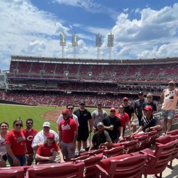 Group of baseball fans in red shirts posing by red stadium seating on a sunny day, with the field in the background.