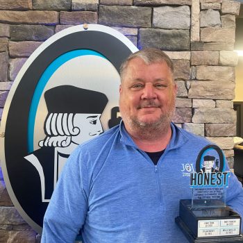 Man in a blue polo shirt poses with an 'Honest' award trophy, in front of a stone wall featuring a large circular logo.