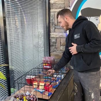 Man in a black hoodie browsing a snack display outside a store, reaching for a candy bar on a wire rack outside the window.