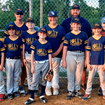 Group of young baseball players and two coaches posing on a dirt field behind a chain-link fence in blue 'Jolly' uniforms and caps.
