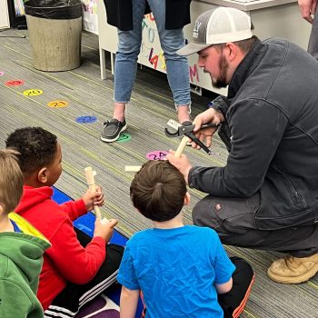 Instructor with a hammer teaches children woodworking with wooden dowels in a classroom.