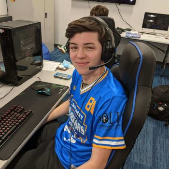 Young man in a blue esports jersey wearing a headset sits at a computer, smiling for the camera in a gaming setup.
