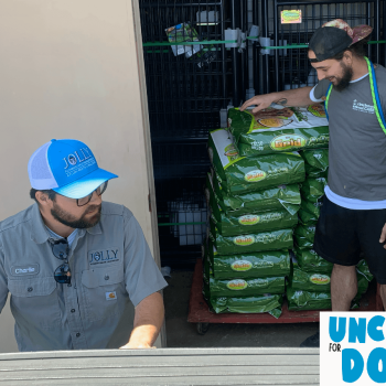 Two volunteers unload large green bags of dog food from a truck; one wears a blue cap and name tag, the other assists from the back of the truck.