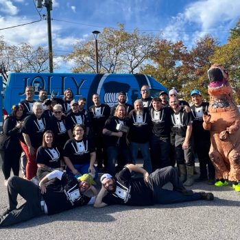 Group of people in matching shirts posing for a team photo in front of a blue van, with a person in a dinosaur costume on the right, outdoors on a sunny day.
