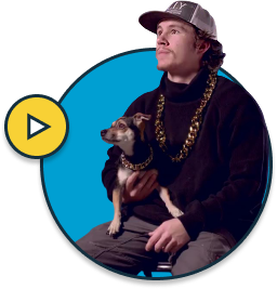 Video thumbnail: man with a cap and braids holding a small dog, yellow play button over a blue circular background, indicating a video.