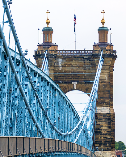 Blue suspension bridge with stone towers and an American flag, seen from the pedestrian walkway along the left side of the structure.