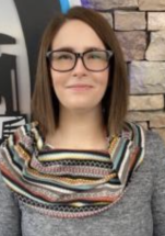Woman with shoulder-length brown hair and black-framed glasses, wearing a patterned scarf, stands in front of a brick wall.