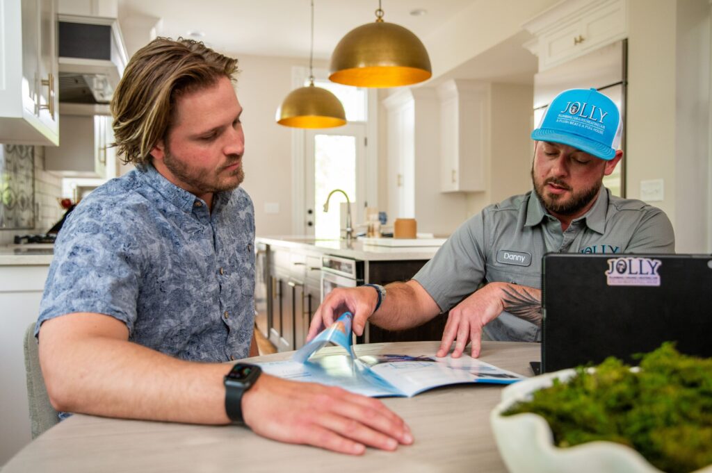 Two men review a brochure at a modern kitchen table, one in a blue patterned shirt and the other in a gray uniform with a blue JOLLY cap, laptop nearby.
