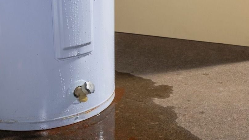 Blue water heater with a leaking inlet valve and a puddle forming on the floor nearby.