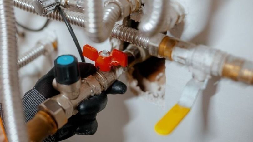 Gloved hand turning a red-handled plumbing shutoff valve under a sink, surrounded by metal pipes and hoses.