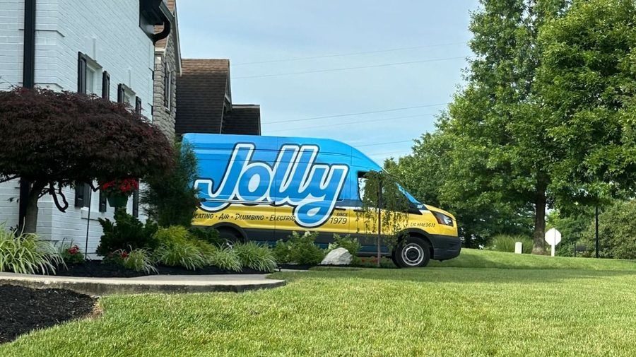 Blue Jolly service van parked on a lawn beside a white house, advertising heating, air, plumbing, and electric services.