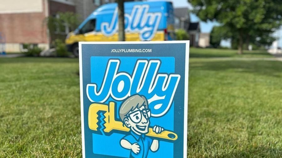 Sign for Jolly Plumbing featuring a cartoon plumber holding a large yellow wrench, with a blue logo on a grassy lawn; van in the background.