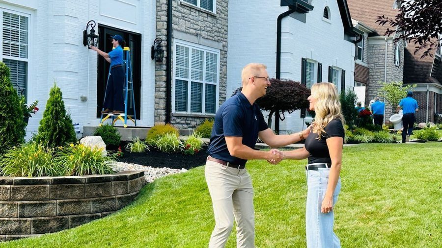 A man and woman shake hands on a green front lawn of a suburban home while a painter on a ladder works on the front door behind them.