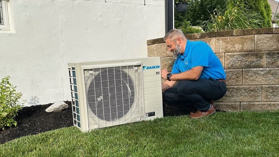 Technician in a blue shirt crouches beside a Daikin outdoor air conditioner unit on a green lawn, inspecting it near a brick wall.