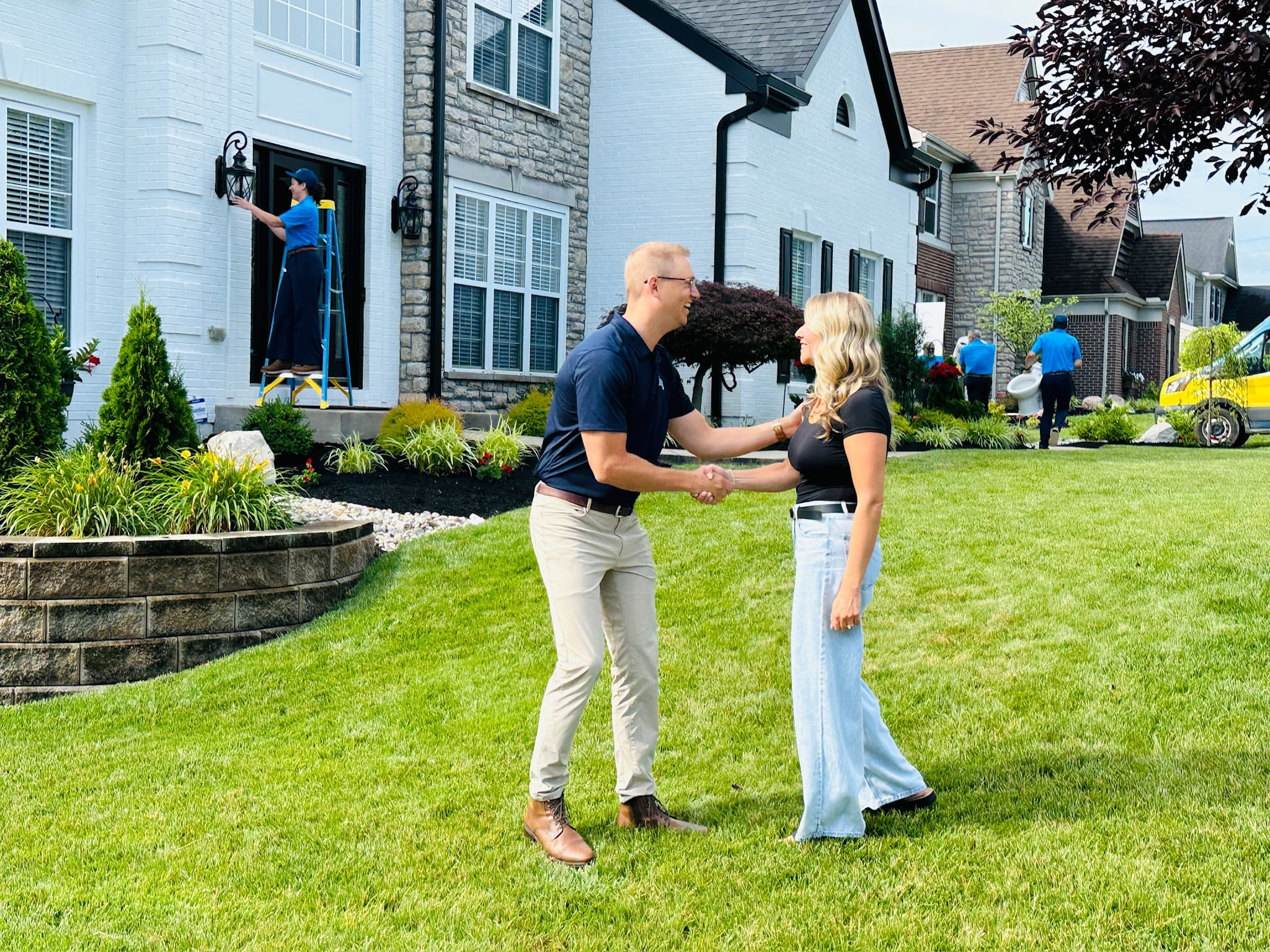 Couple shaking hands on a sunny lawn in front of a white brick house while workers perform exterior maintenance in the background.
