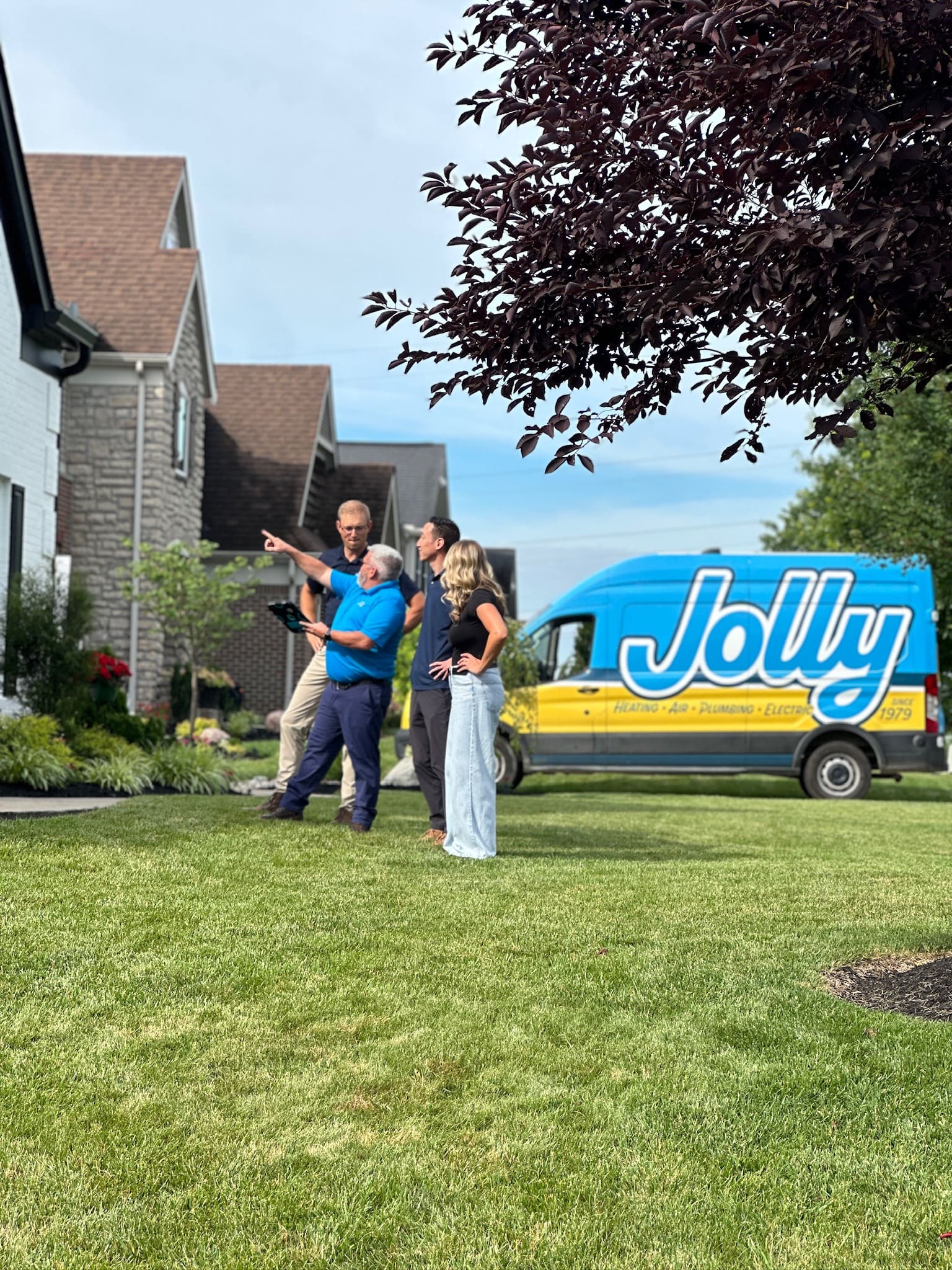 Group of adults standing on a front lawn discussing something, with a blue service van labeled "Jolly" in the background.