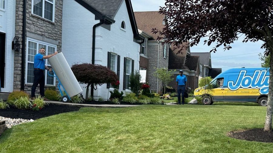 Two workers in blue shirts move a large white appliance on a dolly toward a suburban house with a delivery van parked in the driveway.",