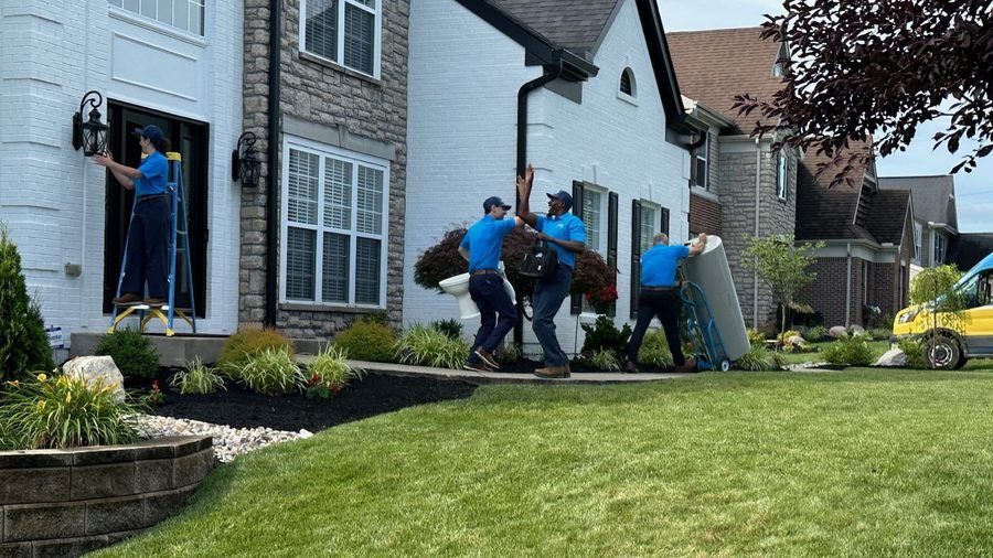 Four movers in blue shirts lift a sofa from a house into a truck on a sunny suburban lawn area.